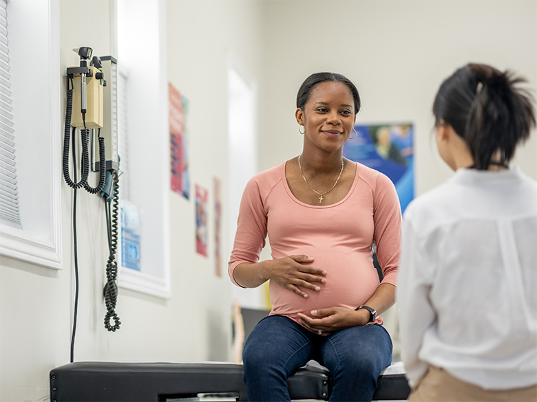 A pregnant woman sits on the bed in an exam room, speaking to her doctor.
