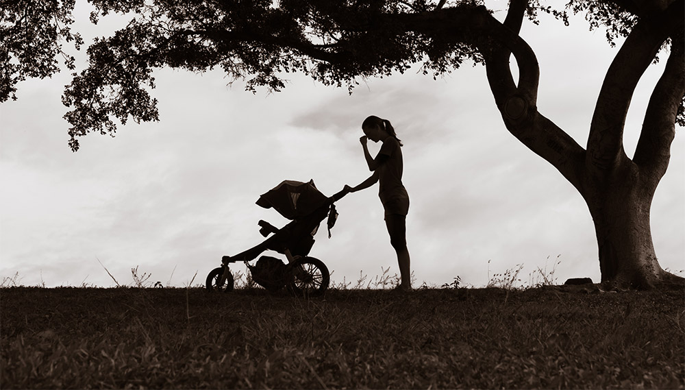 A woman pushing a stroller stands still and holds her hand to her face, as if in emotional pain. 