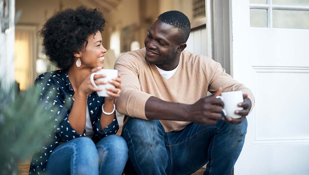 A woman and a man sit on their front step, smiling at each other and holding coffee cups.