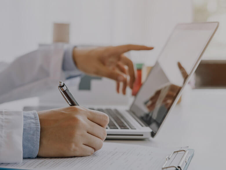 Person in a lab coat writing and pointing at a laptop screen.