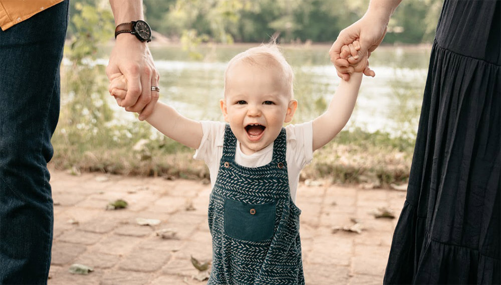 Erica's baby smiles while standing and holding his parents' hands. His parents are on either side of him, out of frame.