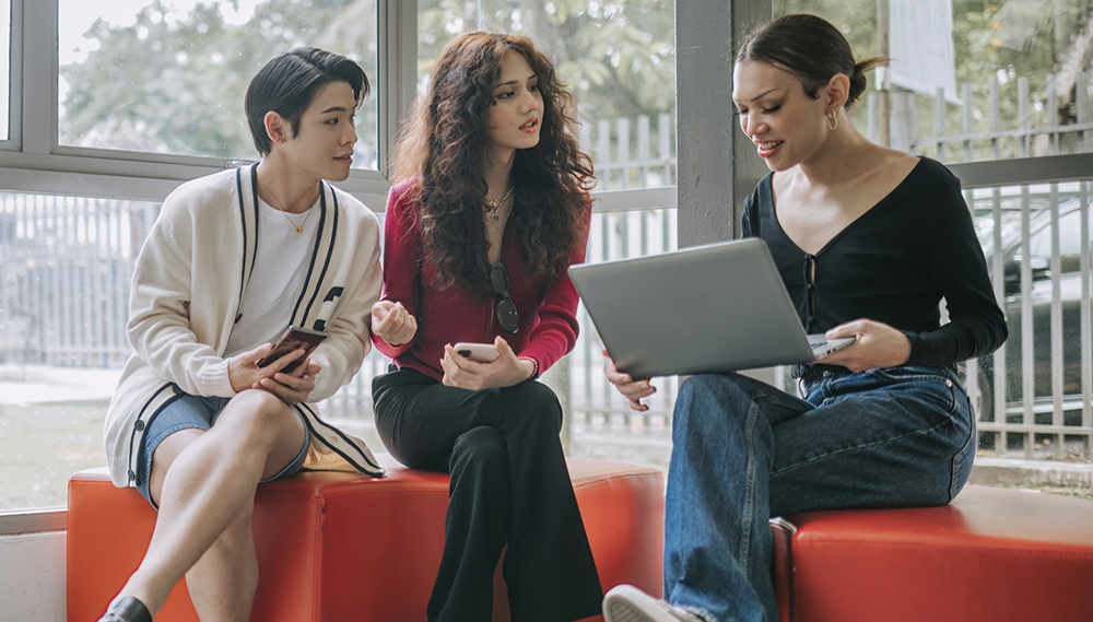 Three students sitting on a bench looking at a laptop.