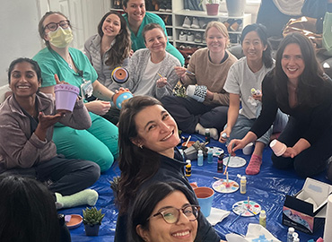 A group of people sit on a tarp painting flower pots together, smiling and showing their work during a creative indoor activity
