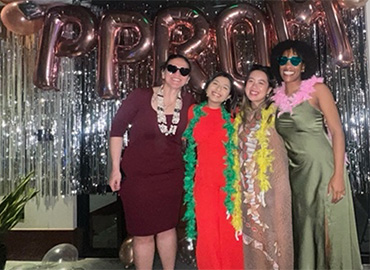 Four women pose in front of silver streamers and balloon letters spelling PPROM, wearing colorful dresses and feather boas.
