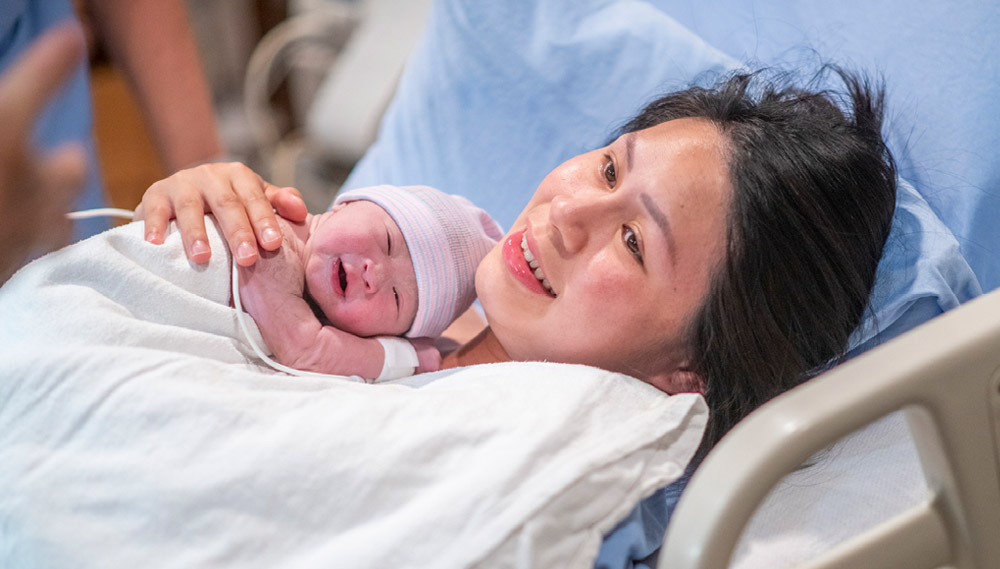 A woman lies in a hospital bed with a newborn baby on her chest.