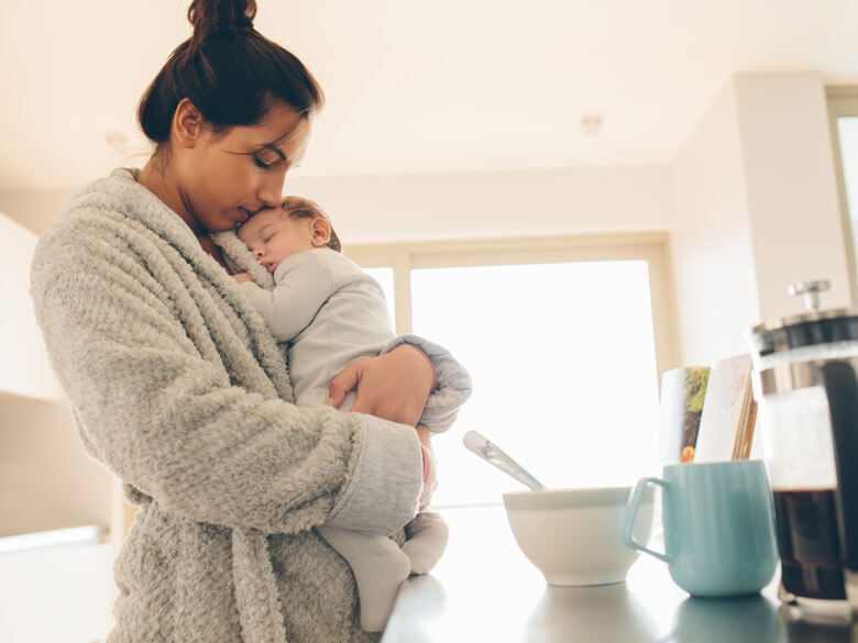 woman in bathrobe cuddling a baby with cereal bowl and coffee cup on a counter